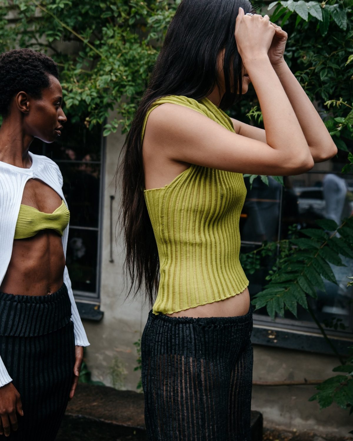 Two women standing outside, one wearing a green pleated top and black sheer knitted skirt and the other wearing a white cut out top, green bralette and black skirt