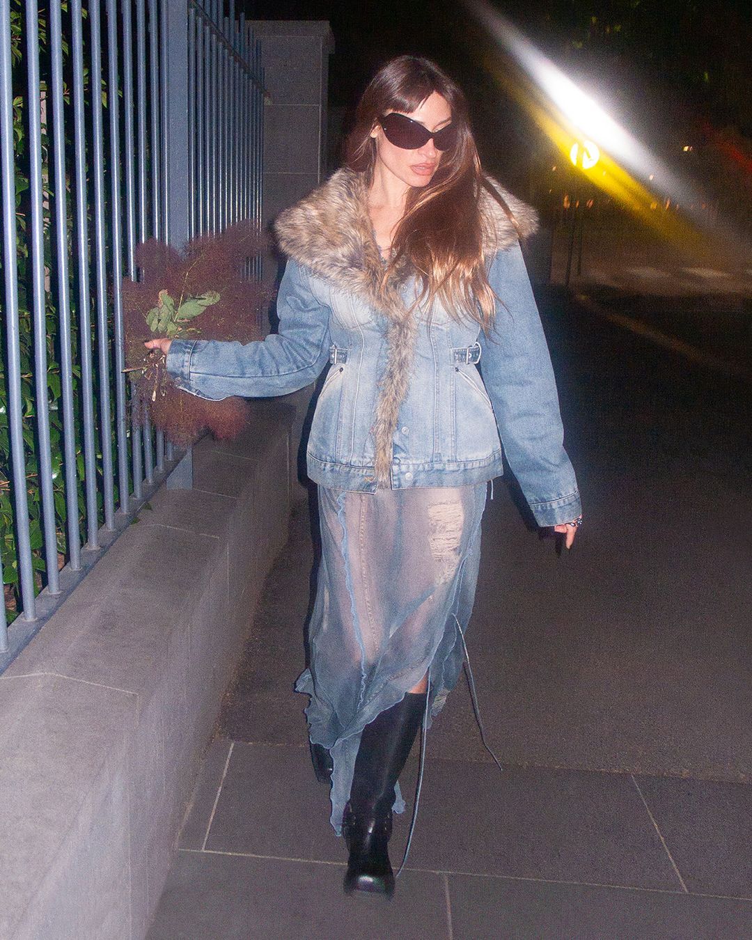 Dark haired woman outside wearing a denim jacket with a faux fur collar, a sheer blue skirt and black knee high boots