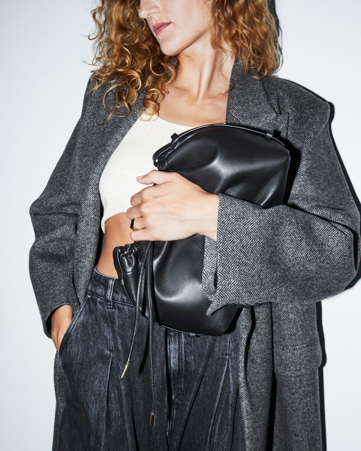 Close up of a woman wearing a long grey blazer, a white tank top, faded black jeans and a black pouch clutch bag