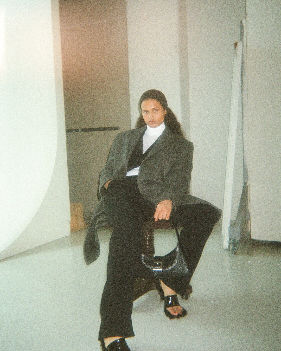 Dark haired woman sitting in a chair wearing a white turtleneck sweater, a black vest, black pants, long grey coat, a black handbag and black slide sandals