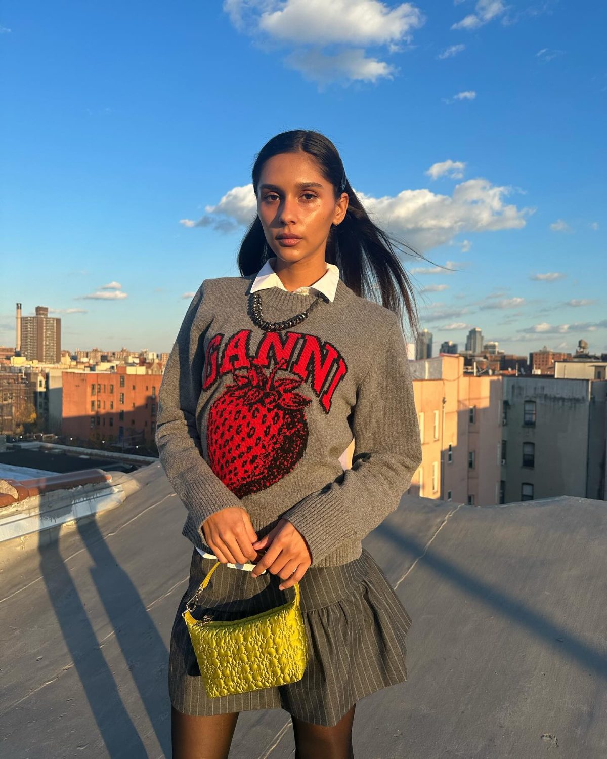 Girl on a rooftop wearing a white button-up shirt, with a grey crewneck sweater with red Ganni text on the front over the top, plus a grey mini skirt