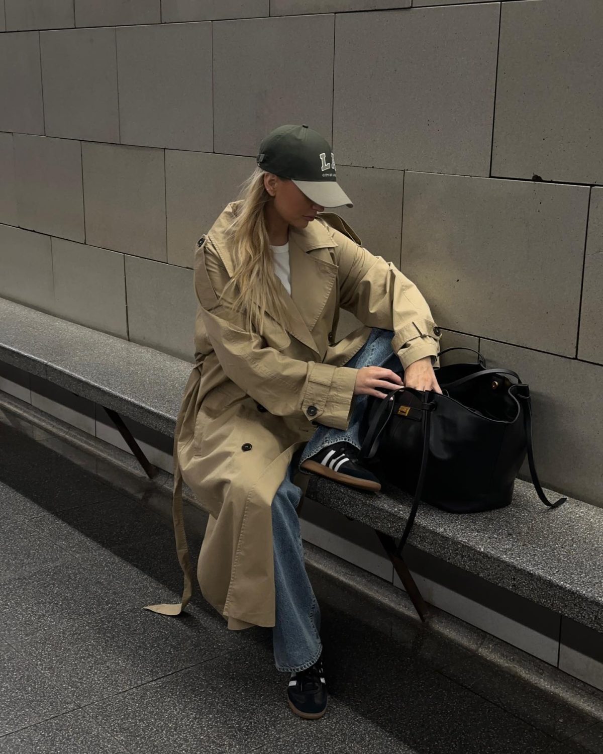 Woman sitting on a grey bench wearing a green cap, a camel trench coat, white top, blue jeans and black sneakers