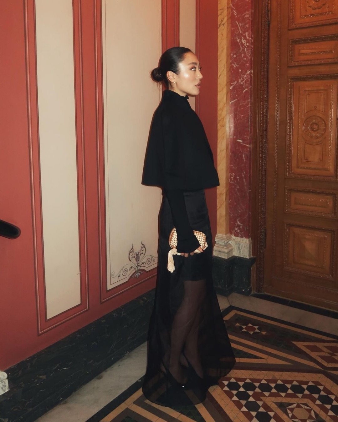 Side view of a woman wearing a black cropped cape with a black sheer maxi skirt layered over a black knee-length skirt, carrying a sparkly clutch