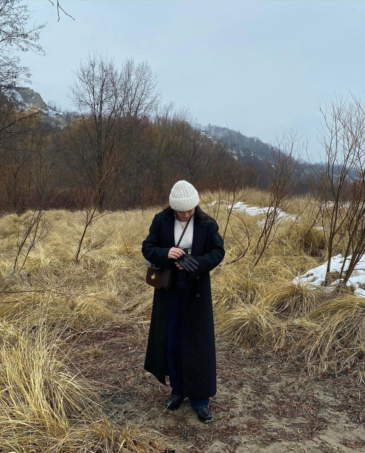 Woman standing in a cold field wearing a long black wool coat over a white top and dark blue jeans, with a white beanie and black boots