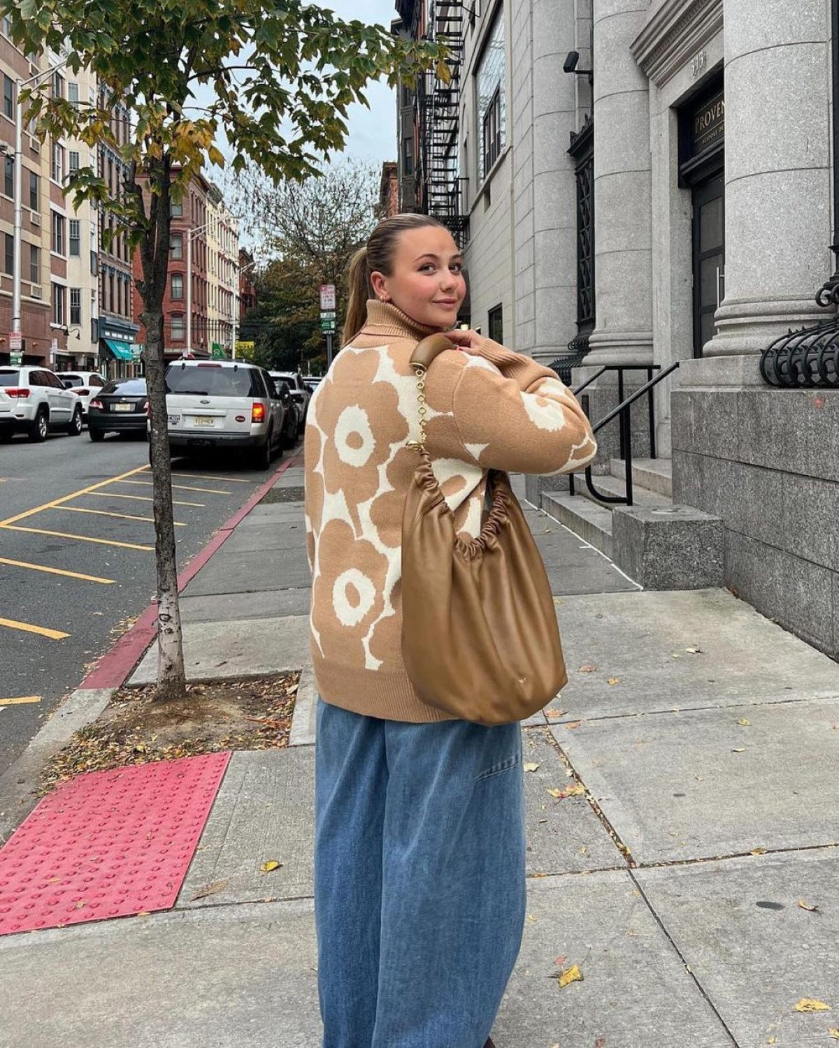 Woman on the sidewalk wearing blue wide leg jeans, a beige and white floral cardigan, and a beige leather shoulder bag