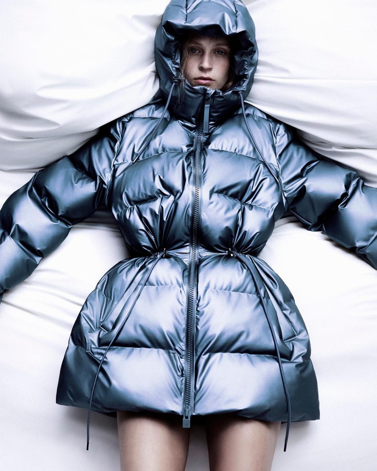 Woman laying in a bed wearing a metallic blue-grey puffer coat with the hood up