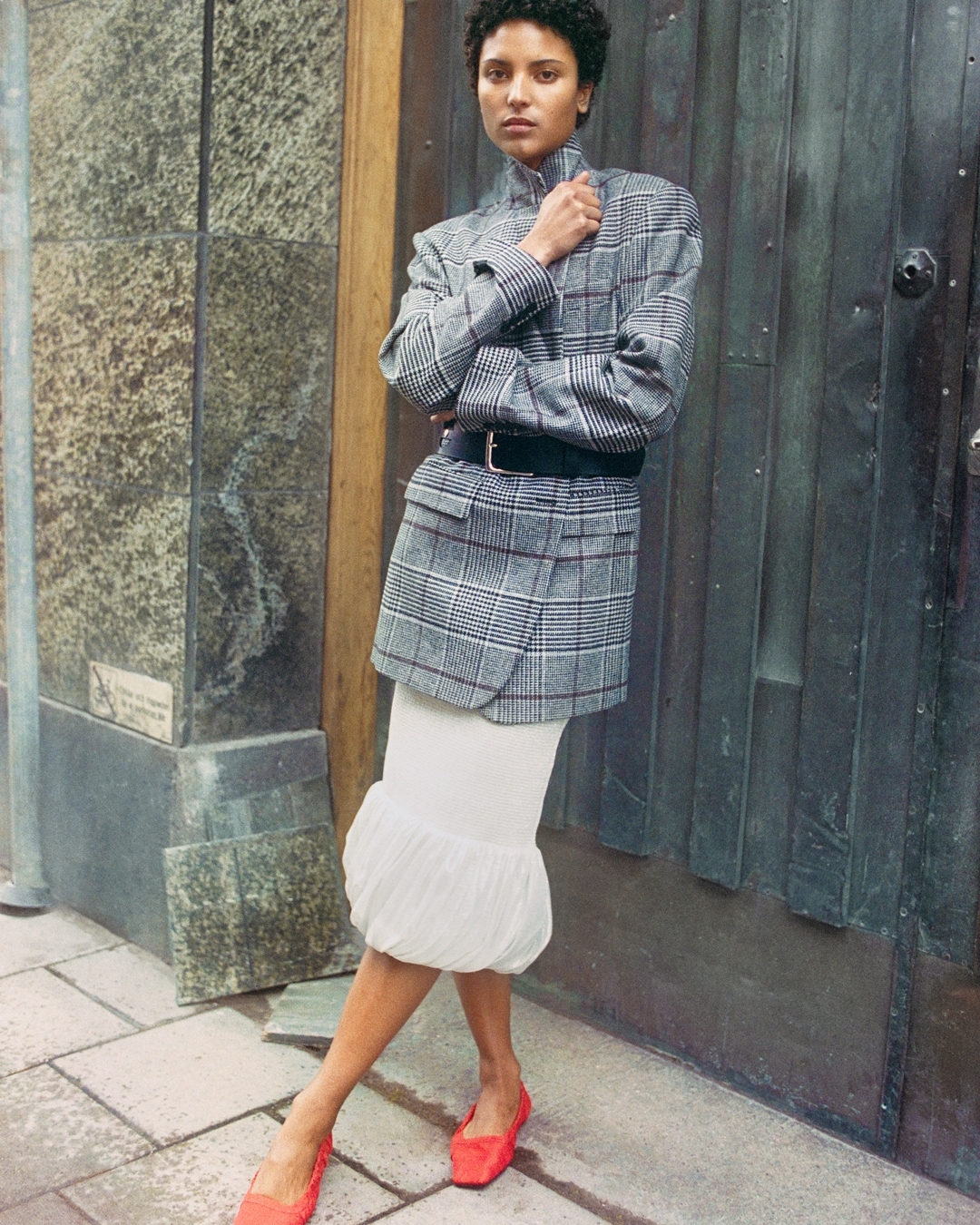 Dark curly haired woman wearing a black plaid blazer belted at the waist a white midi skirt and orange ballet flats