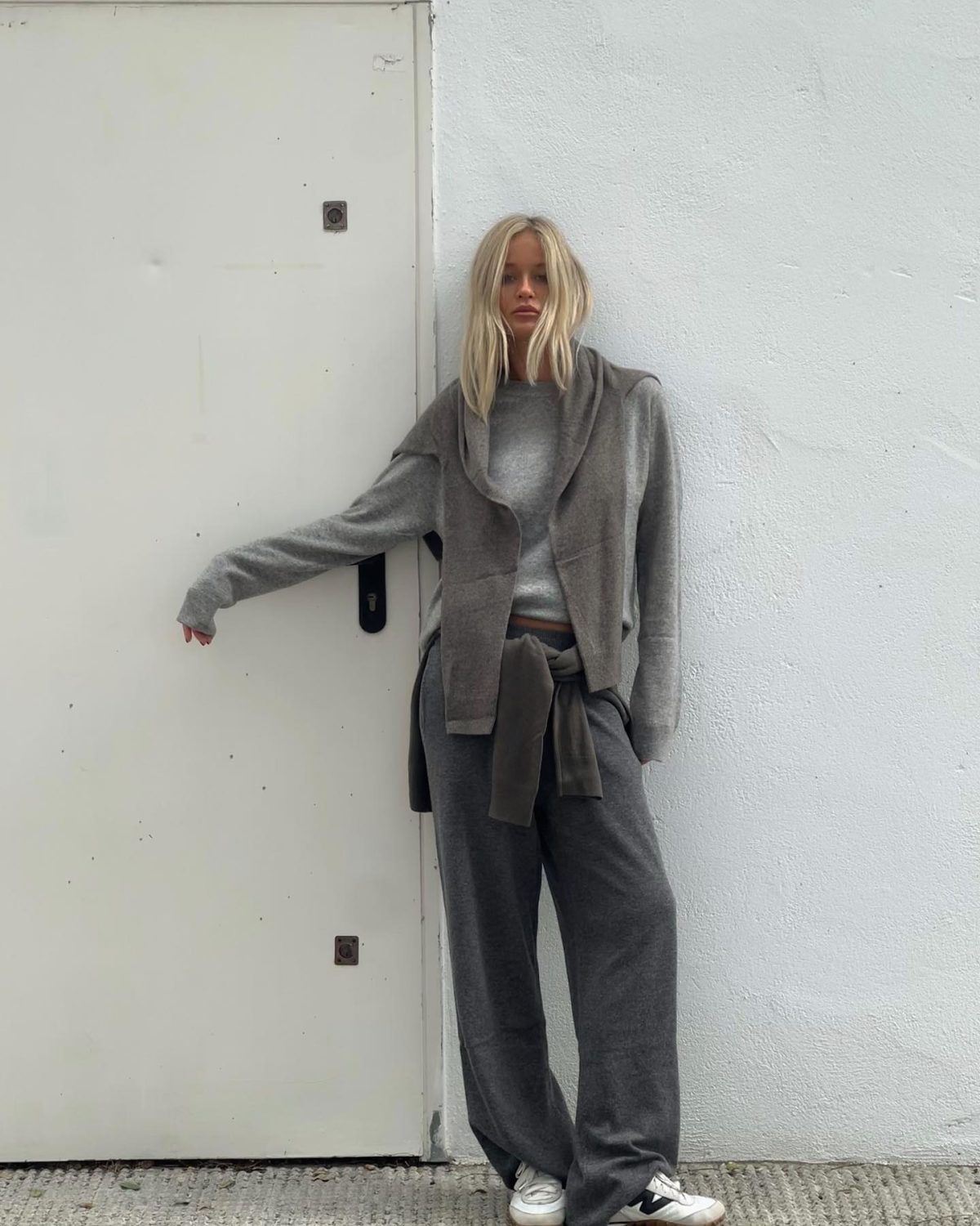 Blonde woman wearing a grey sweater a darker grey cardigan over her shoulder dark grey baggy pants and white sneakers